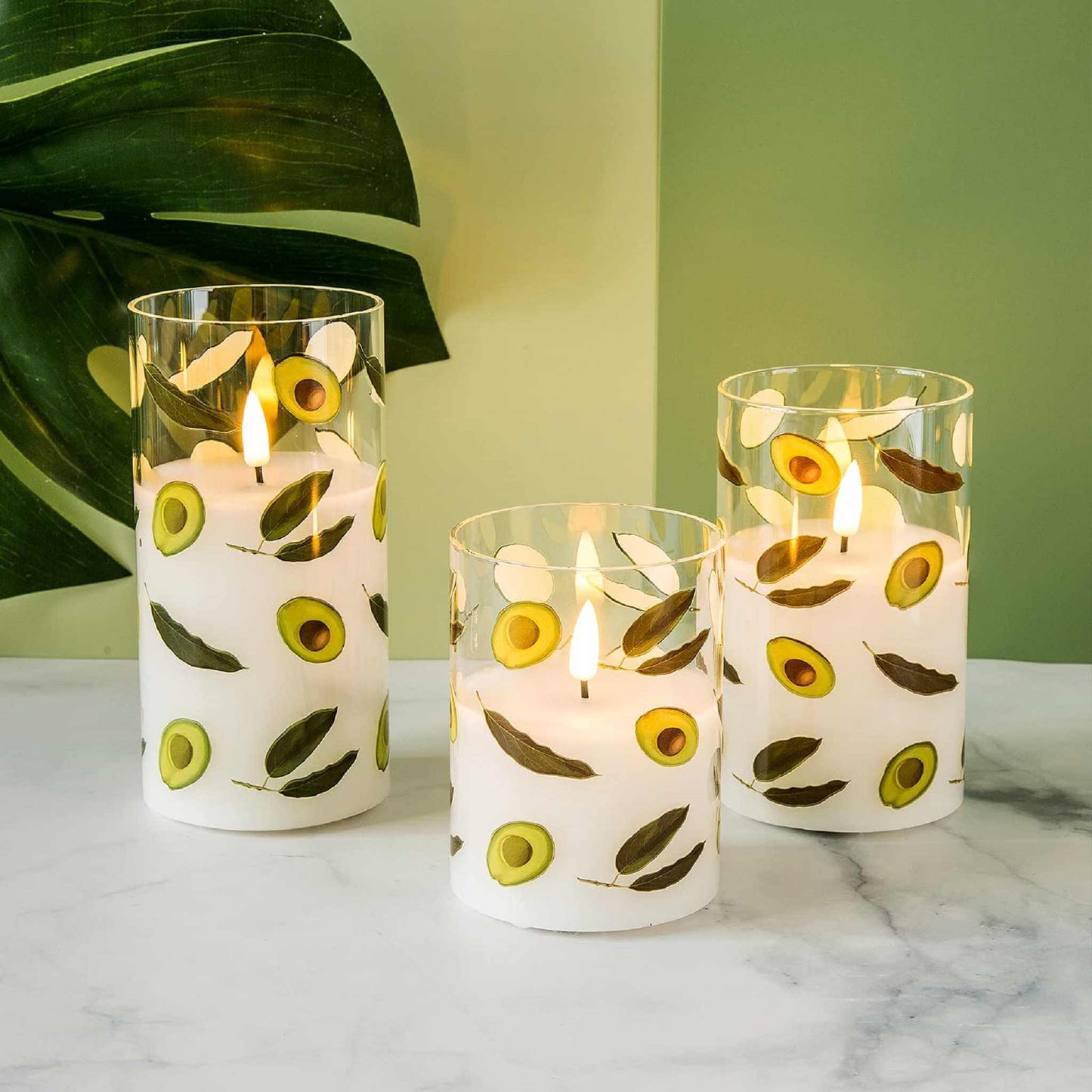 Three lit avocado glass flameless candles placed on a dining table with a green wall and plants in the background