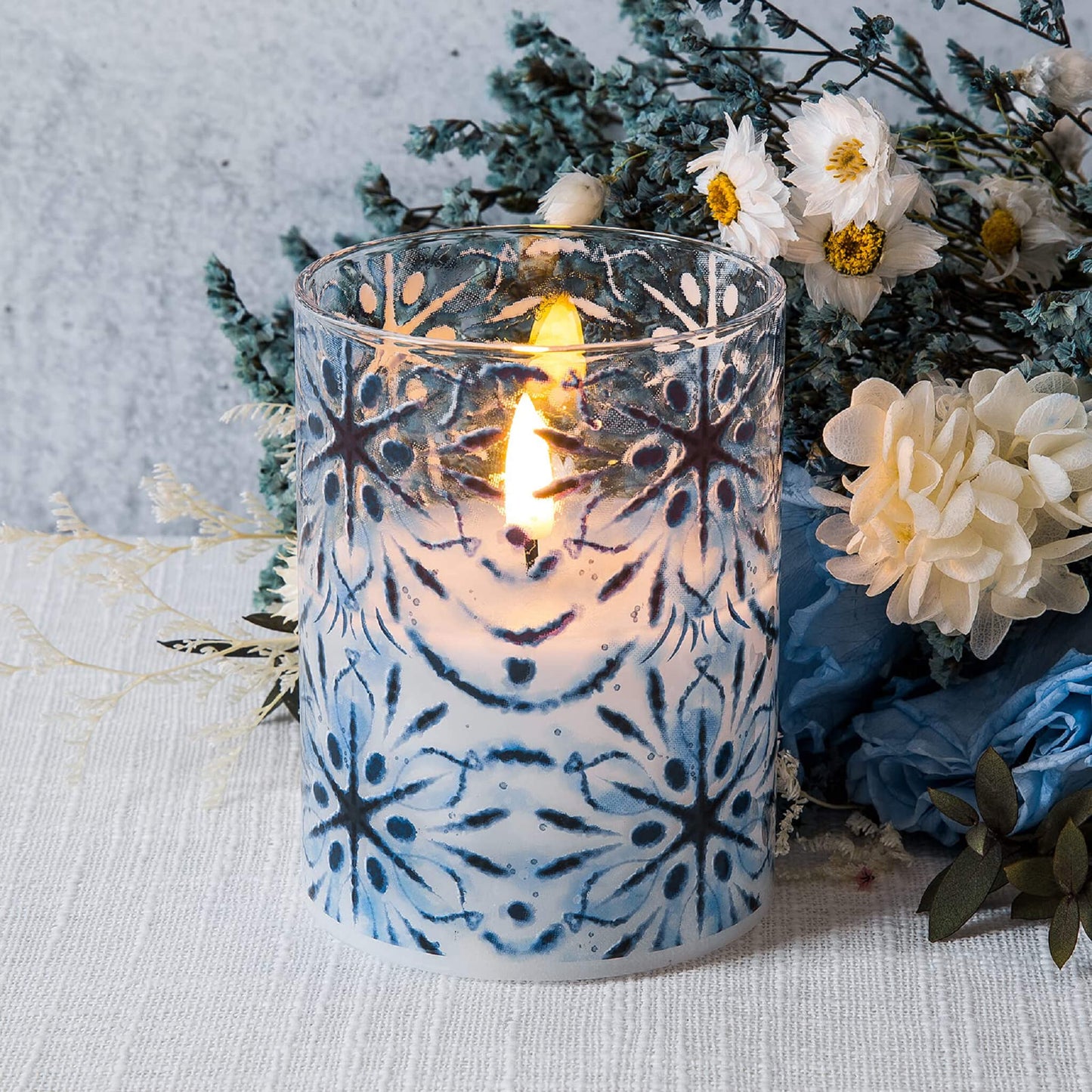 one blue snowflakes glass flameless candle placed on a white tablecloth, next to a bouquet, white blue flowers and some green plants
