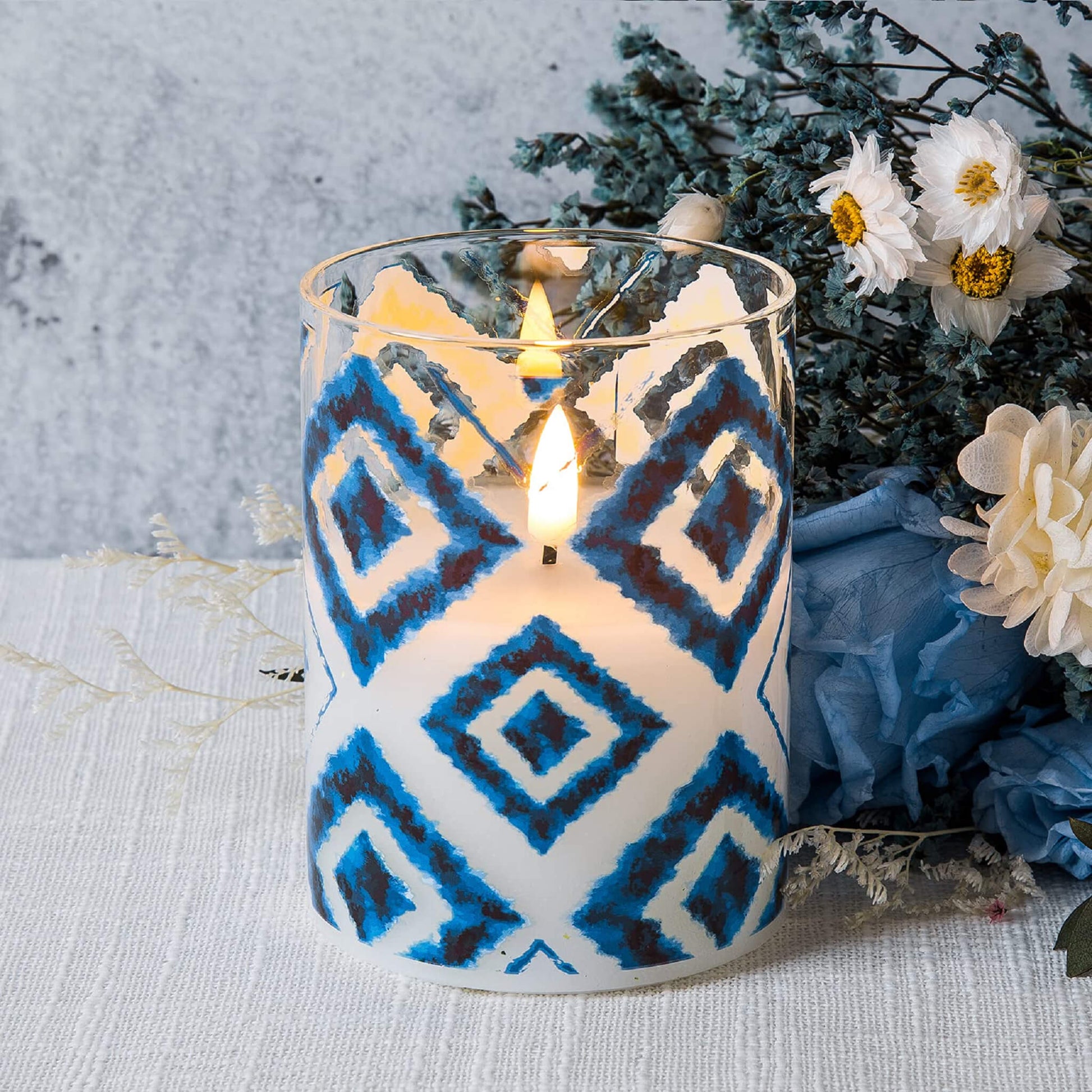 A lit Flickering Blue White Geometric led candle lies on a white tablecloth with a bouquet of flowers behind it