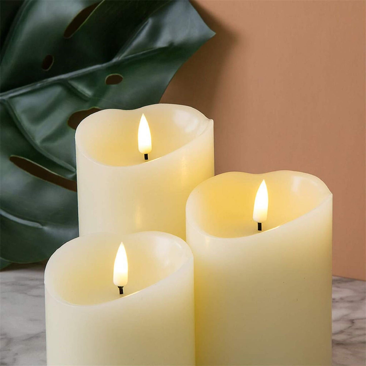 3 led ivory timer flameless pillar candles on a marble table top with a huge green leaf behind