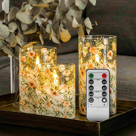 A set of 3 eywamage floral glass flameless candles and a remote on a plate on a table with a bouquet of greenery behind