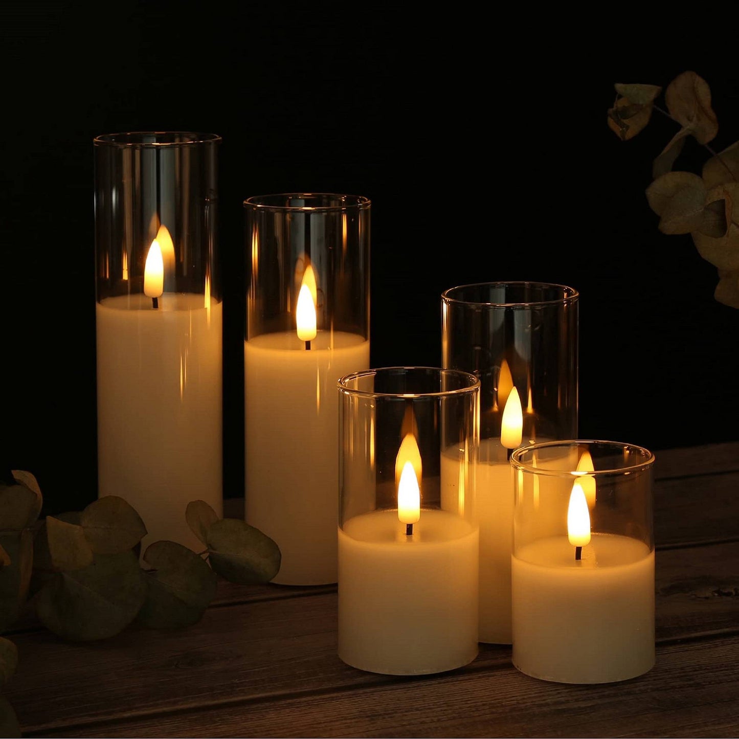 A set of five flameless clear glass candles of different lengths and some greenery on a table
