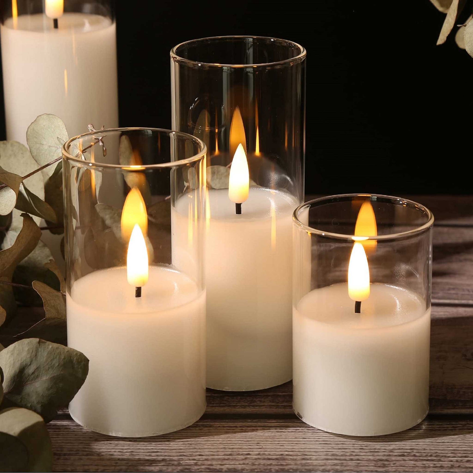 Four slim clear glass flameless candles on a table with green plants