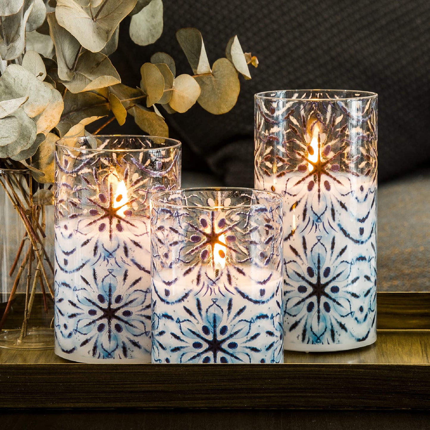 three blue snowflakes glass flameless candles light up on a wooden square plate with a transparent vase next to it with some green foliage