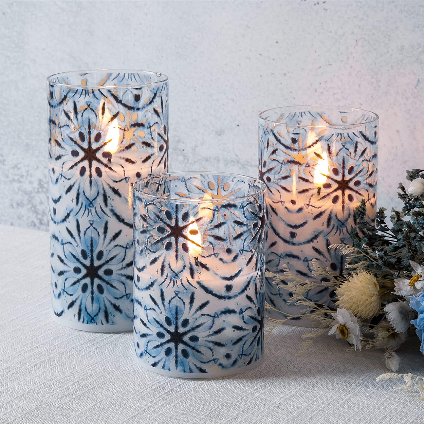 three blue snowflakes glass flameless candles placed on a white tablecloth, next to a bouquet, white blue flowers and some green plants