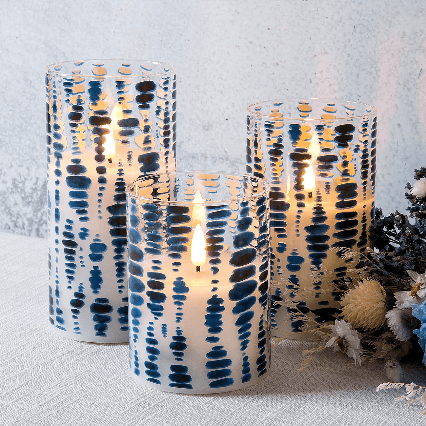 Three blue dots glass flameless candles are lit and placed on a white tablecloth with some plants beside them including several white and blue flowers