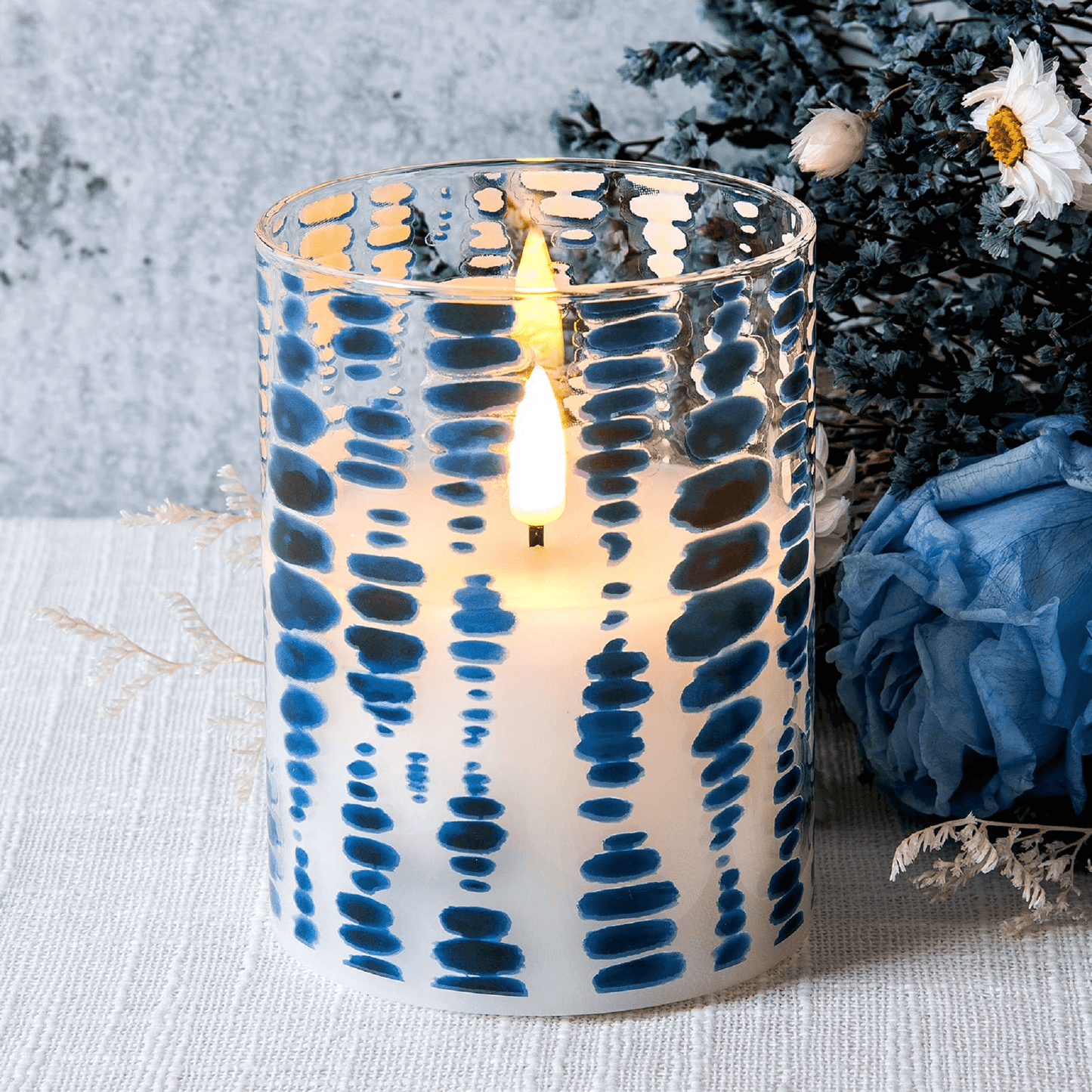A blue dots glass flameless candle lit and placed on a white tablecloth with some greenery and a blue flower in the background