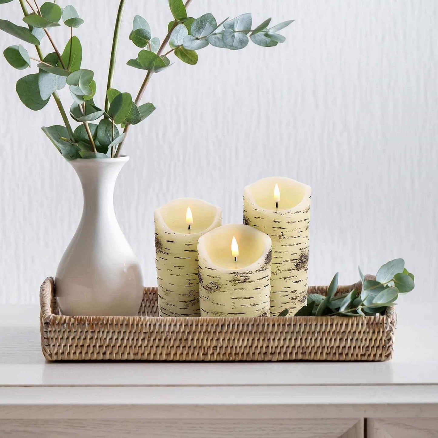 A bamboo plate with three melted top birch pattern pillar candles and a vase with some greenery on the dining table