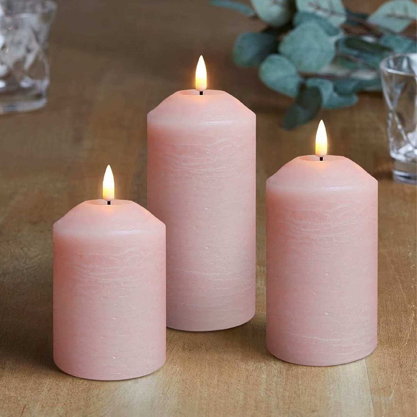 There are three blush pink flameless pillar candles, two glasses and some green plants on the table