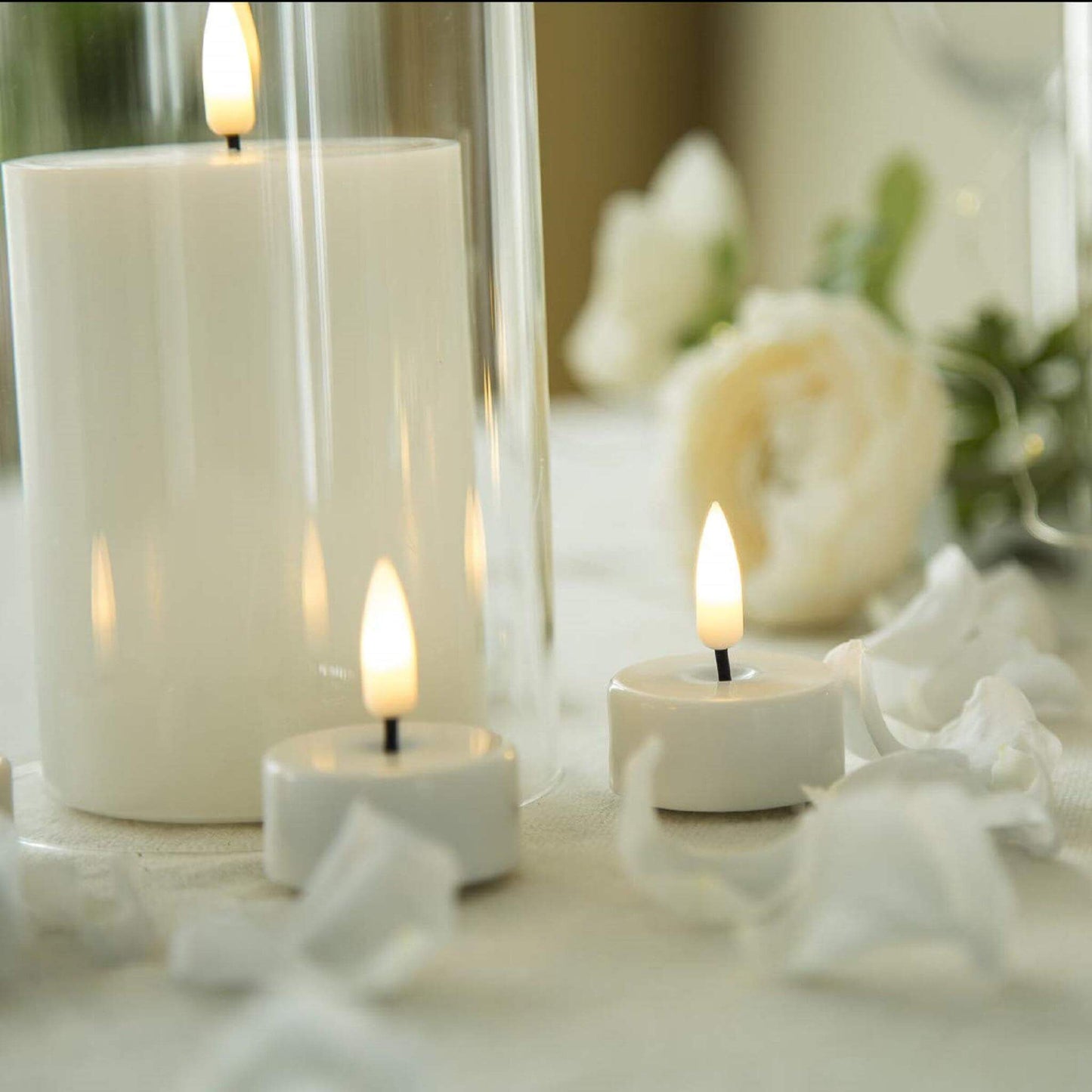 Two electronic tea lights, a glass and white flower petals are on the table. There is a pillar flameless candle in the glass.