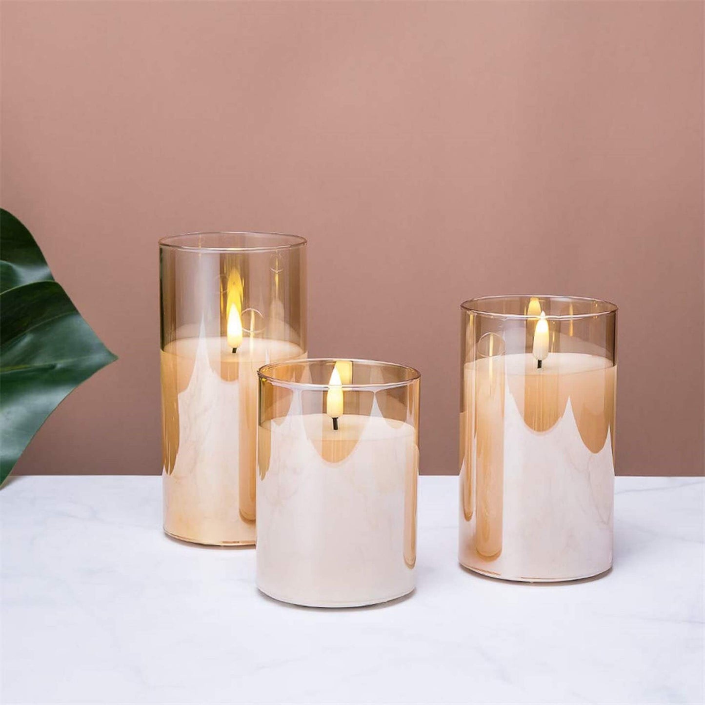 Three eywamage gold glass flameless candles with a remote on a white table with an orange wall and a large green leaf behind