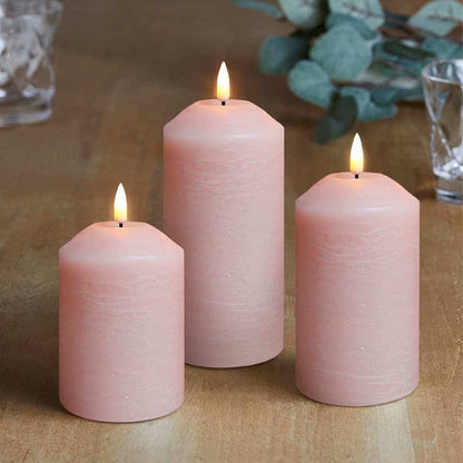 There are three blush pink flameless pillar candles, two glasses and some green plants on the table